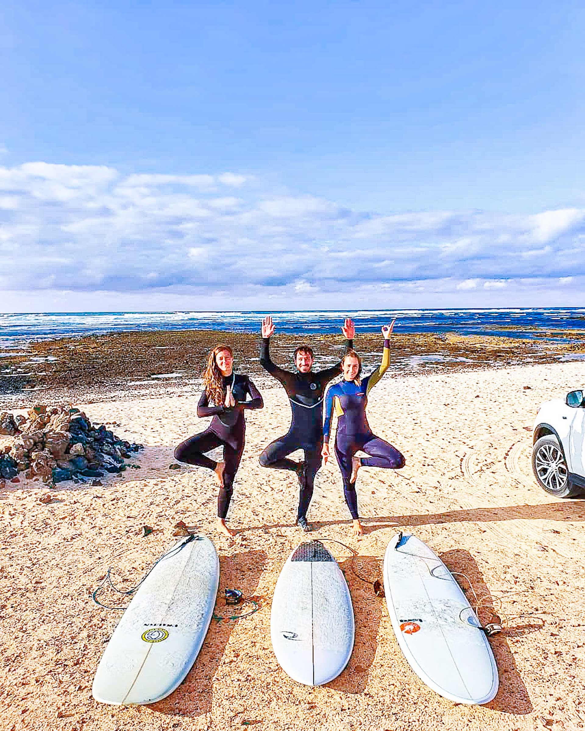 Three young people practising the yoga Tree Pose in wetsuits with surfboards in Fuerteventura